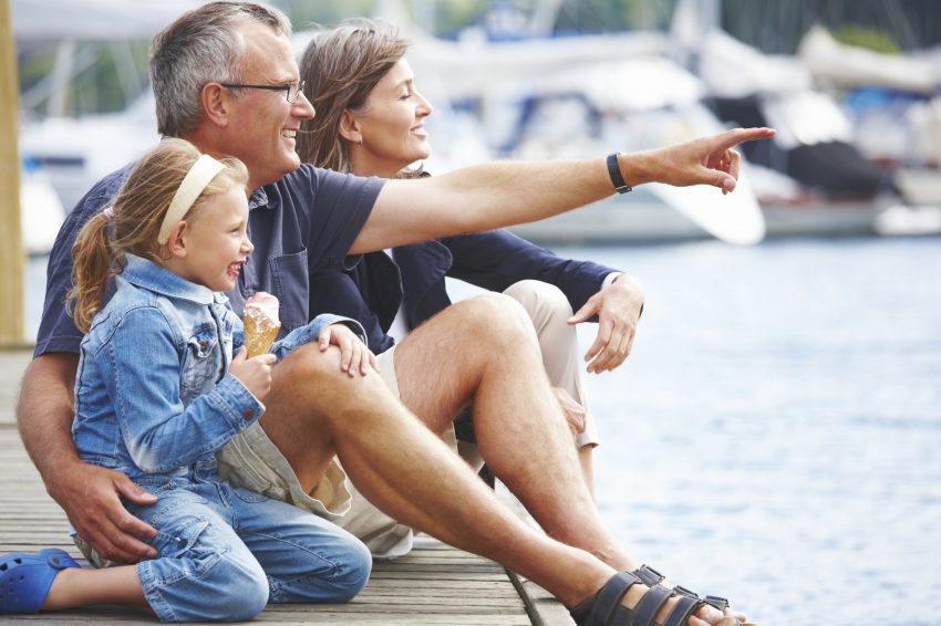 Happy Grandparents sitting with their granddaughter on a pier, pointing out at the boats - copyspace